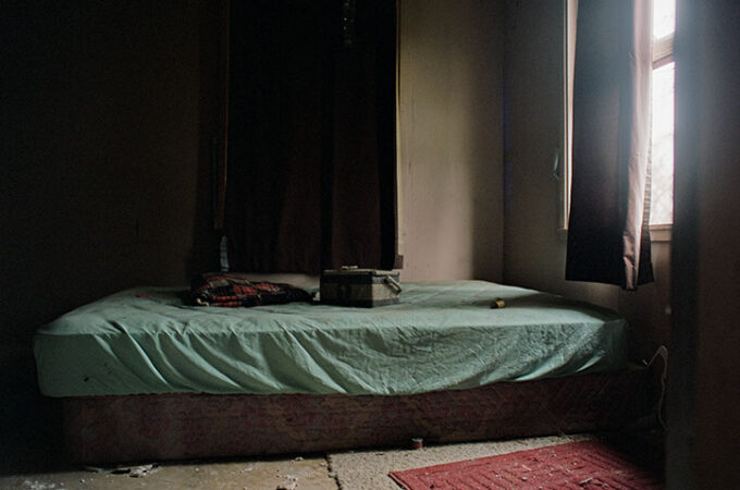 An abandoned room at Lily of the Valley Campground with a bed, light green sheet, dark curtains, and a sunlit window.