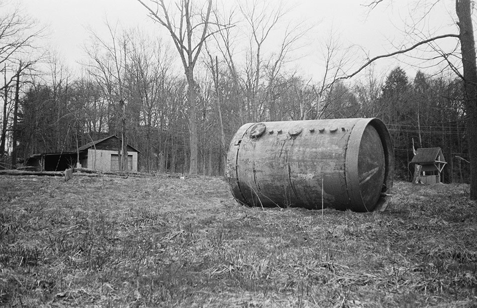 Rusted tank on bare ground, with abandoned structures and leafless trees at Lily of the Valley Campground, Thompson,