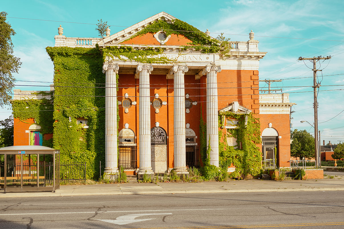 An abandoned brick bank building, Lake Shore Bank, covered in green ivy, with four white columns under a clear blue sky.