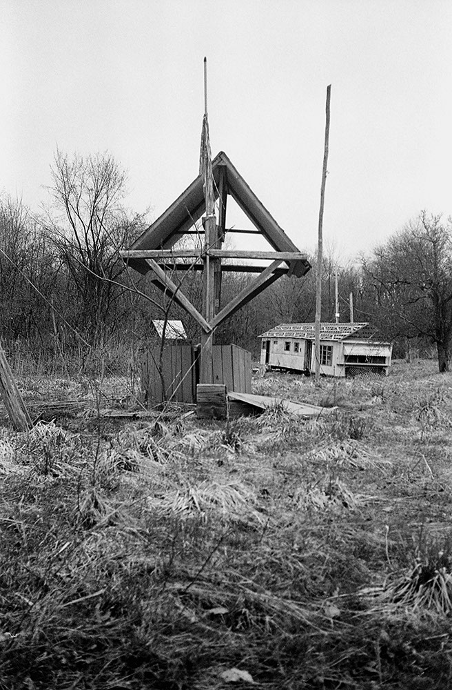 Monochrome view of Lily of the Valley Campground showing a weathered wooden arch and a decaying building in dry grass.